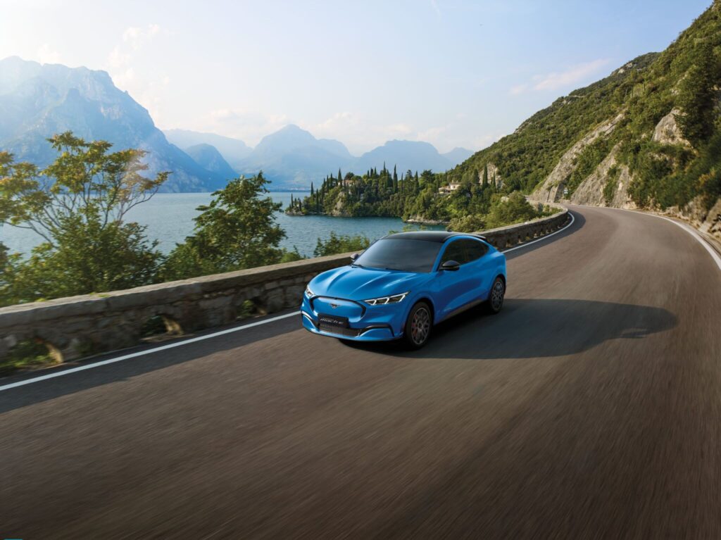 Blue Ford Mustang Mach-E driving along a scenic lakeside mountain road under clear skies.