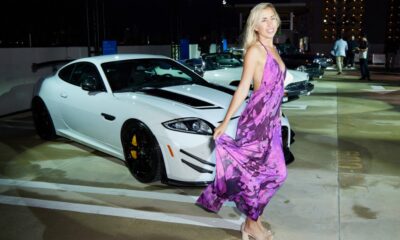 A woman in a flowing purple dress poses in front of a white Jaguar sports car at an evening outdoor event.