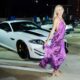 A woman in a flowing purple dress poses in front of a white Jaguar sports car at an evening outdoor event.