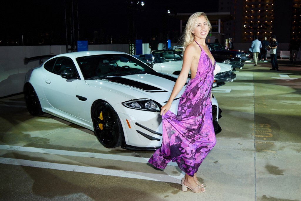 A woman in a flowing purple dress poses in front of a white Jaguar sports car at an evening outdoor event.
