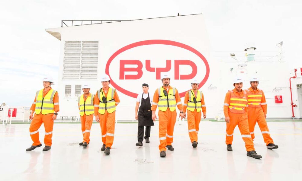 A group of Filipino seafarers and crew members in orange coveralls and safety vests walk on the deck of a BYD roll-on/roll-off vessel, with the large BYD logo visible in the background.