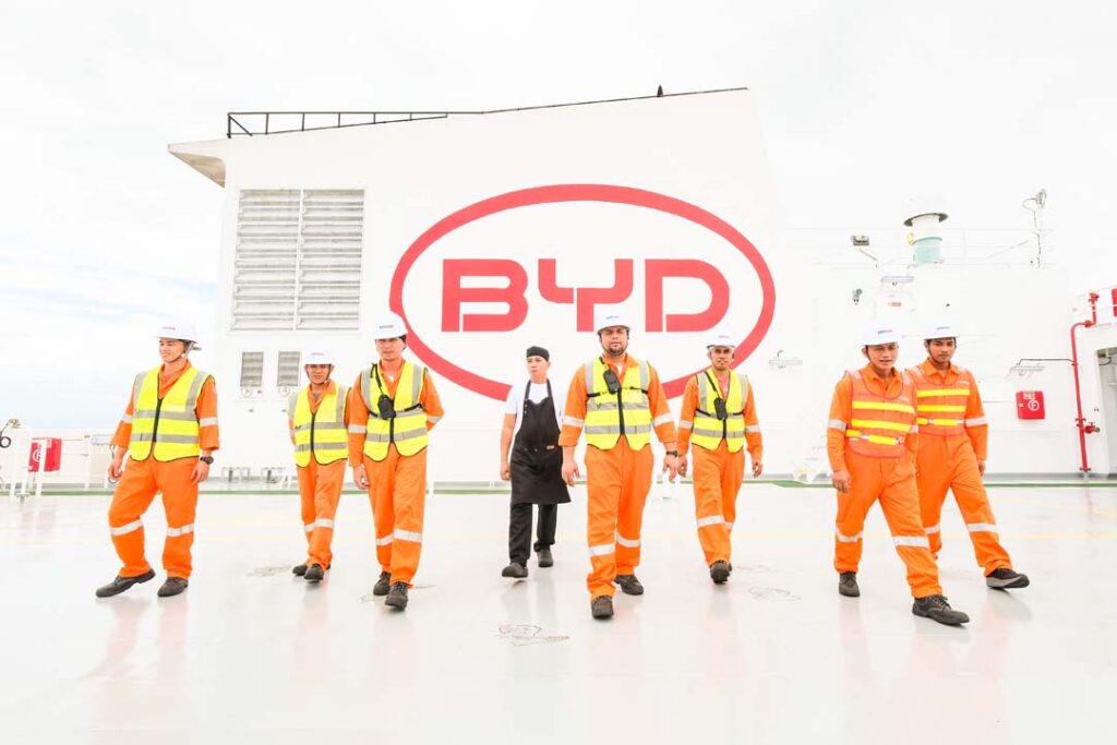 A group of Filipino seafarers and crew members in orange coveralls and safety vests walk on the deck of a BYD roll-on/roll-off vessel, with the large BYD logo visible in the background.