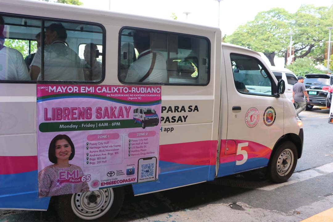 A white and pink Toyota Lite Ace community shuttle in Pasay City with a poster advertising “Libreng Sakay” (Free Ride) from Monday to Friday, 6 a.m. to 6 p.m., featuring Mayor Emi Calixto-Rubiano and the list of shuttle zones and stops.