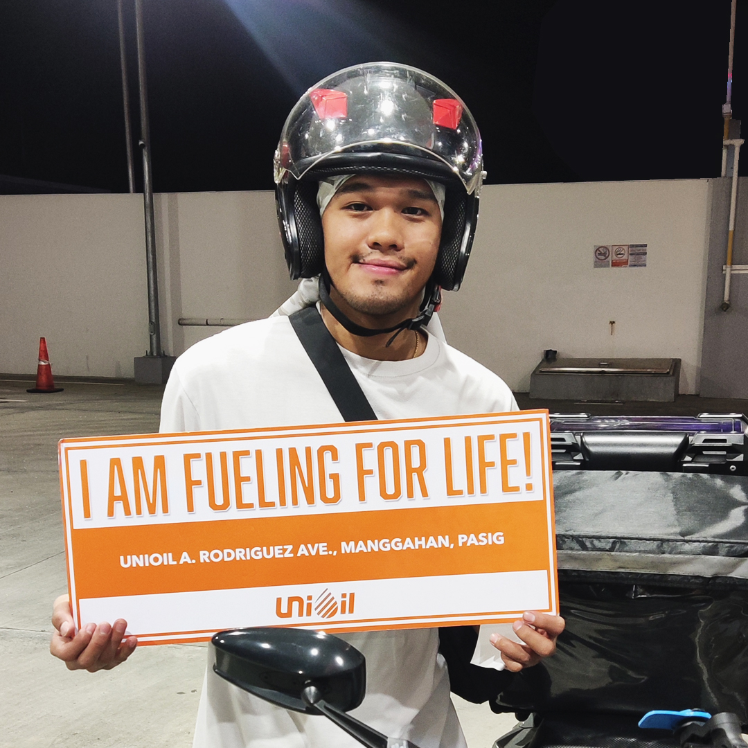 A smiling motorcycle rider wearing a helmet holds a sign that reads "I AM FUELING FOR LIFE!" at a Unioil station in Pasig.