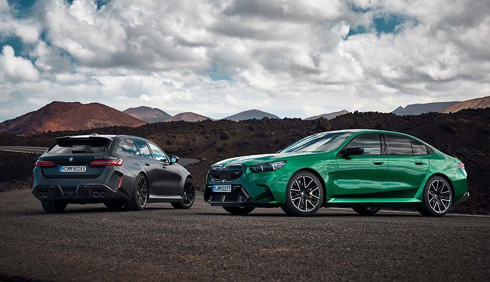 The 2025 BMW M5 in bright green (sedan) and matte black (Touring wagon) parked side by side on a volcanic mountain road under dramatic clouds.