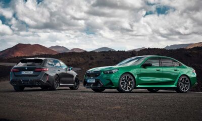 The 2025 BMW M5 in bright green (sedan) and matte black (Touring wagon) parked side by side on a volcanic mountain road under dramatic clouds.