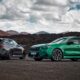 The 2025 BMW M5 in bright green (sedan) and matte black (Touring wagon) parked side by side on a volcanic mountain road under dramatic clouds.