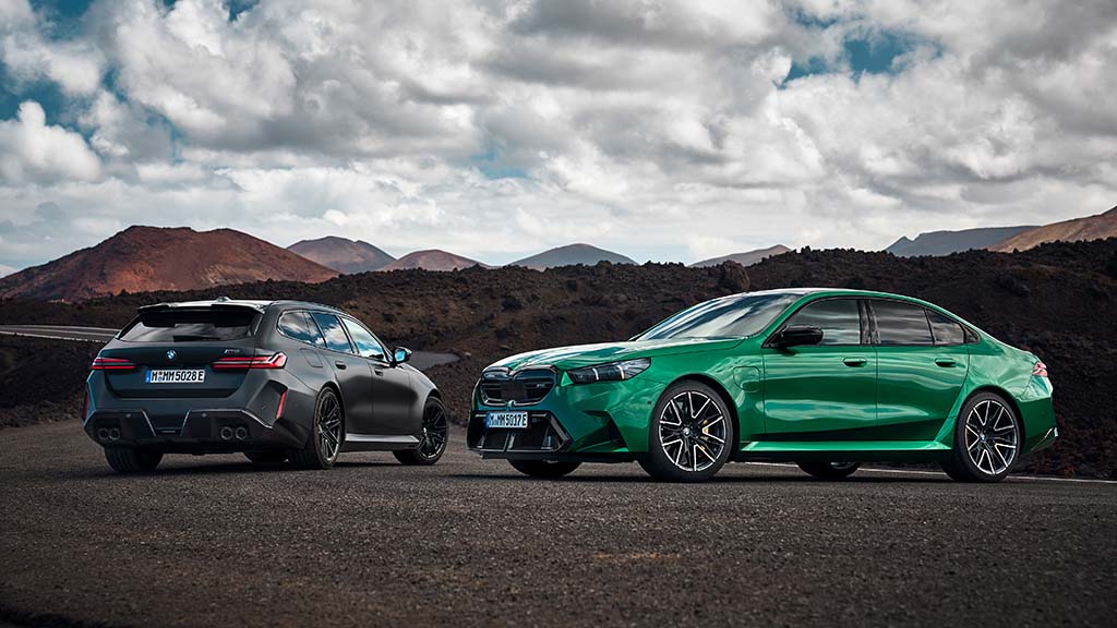 The 2025 BMW M5 in bright green (sedan) and matte black (Touring wagon) parked side by side on a volcanic mountain road under dramatic clouds.