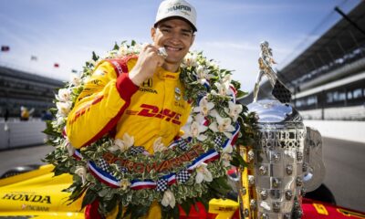 Alex Palou celebrates in victory lane wearing a champion’s cap and floral wreath, standing beside the Borg-Warner Trophy after winning the Indianapolis 500.