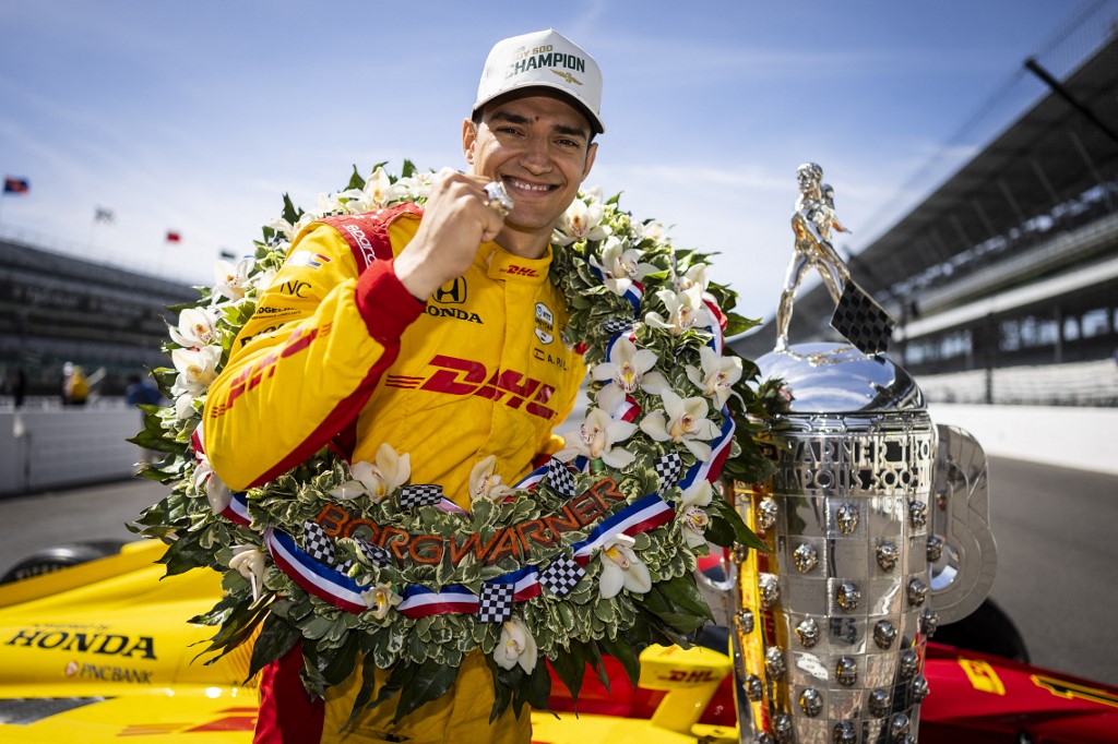 Alex Palou celebrates in victory lane wearing a championâs cap and floral wreath, standing beside the Borg-Warner Trophy after winning the Indianapolis 500.