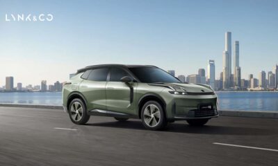 Lynk & Co 08 EM-P plug-in hybrid SUV in Mineral Green driving on a city road with skyline and waterfront in the background.