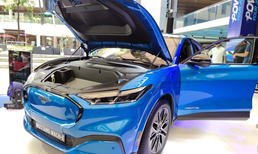 A bright blue Ford Mustang Mach-E electric SUV on display at an outdoor mall event, with its hood open to reveal the front trunk, and both front doors open for viewing.