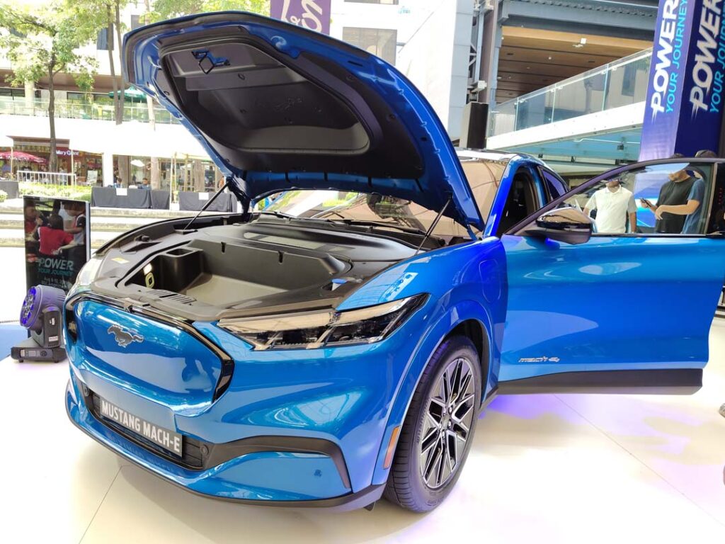 A bright blue Ford Mustang Mach-E electric SUV on display at an outdoor mall event, with its hood open to reveal the front trunk, and both front doors open for viewing.