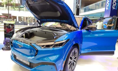 A bright blue Ford Mustang Mach-E electric SUV on display at an outdoor mall event, with its hood open to reveal the front trunk, and both front doors open for viewing.