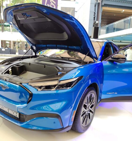 A bright blue Ford Mustang Mach-E electric SUV on display at an outdoor mall event, with its hood open to reveal the front trunk, and both front doors open for viewing.