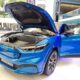A bright blue Ford Mustang Mach-E electric SUV on display at an outdoor mall event, with its hood open to reveal the front trunk, and both front doors open for viewing.