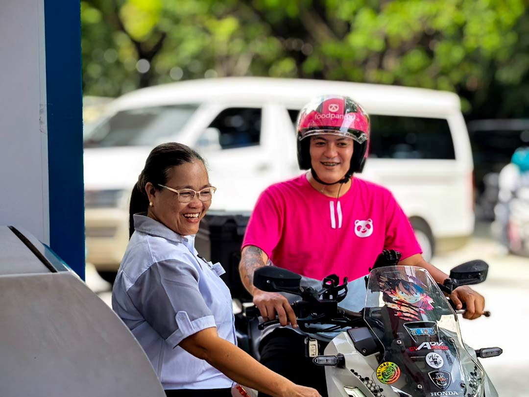A smiling fuel station attendant assists a foodpanda rider on a motorcycle at a Caltex station.