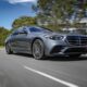 A silver Mercedes-Benz luxury sedan driving on a sunny road, captured from a low front-side angle.