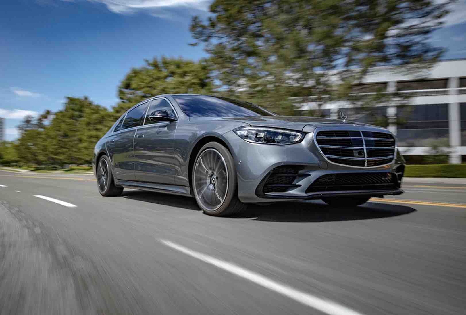 A silver Mercedes-Benz luxury sedan driving on a sunny road, captured from a low front-side angle.