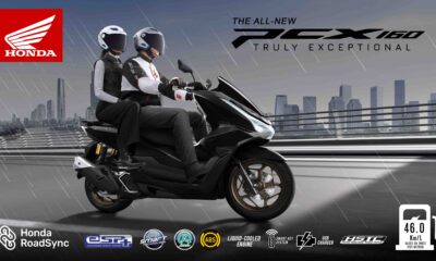 Two riders on a black Honda PCX160 scooter drive confidently in the rain across a modern city bridge, with visible rain streaks and city skyline in the background.