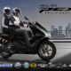 Two riders on a black Honda PCX160 scooter drive confidently in the rain across a modern city bridge, with visible rain streaks and city skyline in the background.