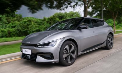 Kia EV6 electric vehicle driving along a tree-lined road, showcasing sleek design and aerodynamic curves under overcast skies.