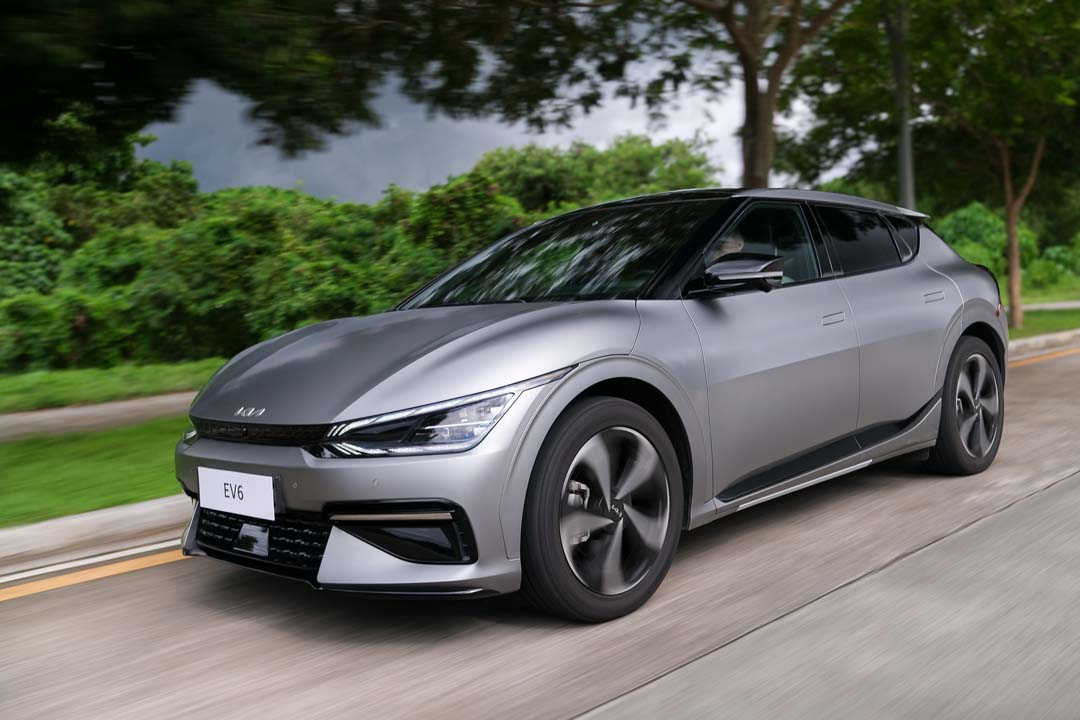 Kia EV6 electric vehicle driving along a tree-lined road, showcasing sleek design and aerodynamic curves under overcast skies.