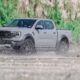 A Ford Ranger Raptor 3.0L V6 drives through a muddy off-road trail, splashing water as it powers forward.