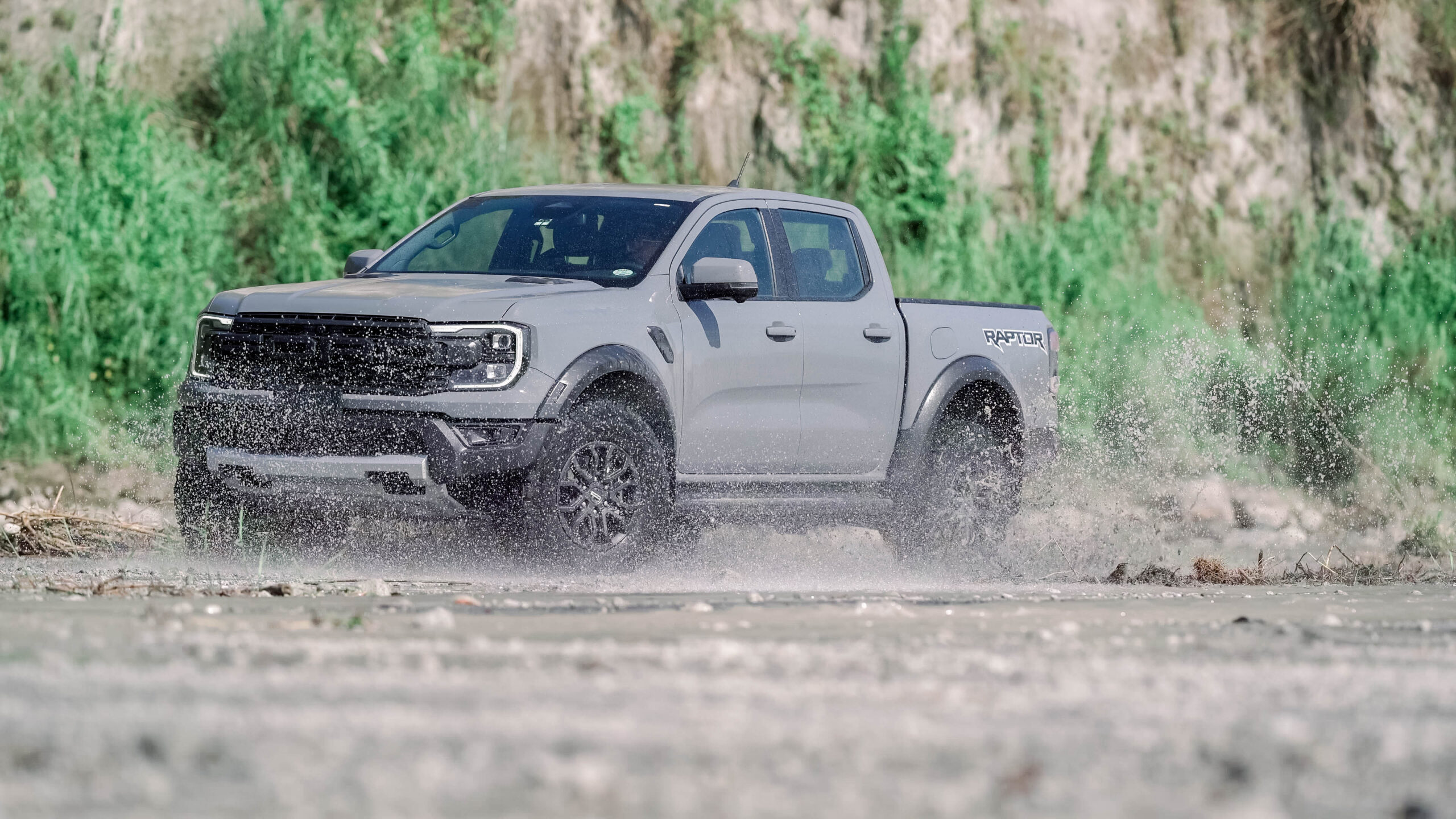 A Ford Ranger Raptor 3.0L V6 drives through a muddy off-road trail, splashing water as it powers forward.