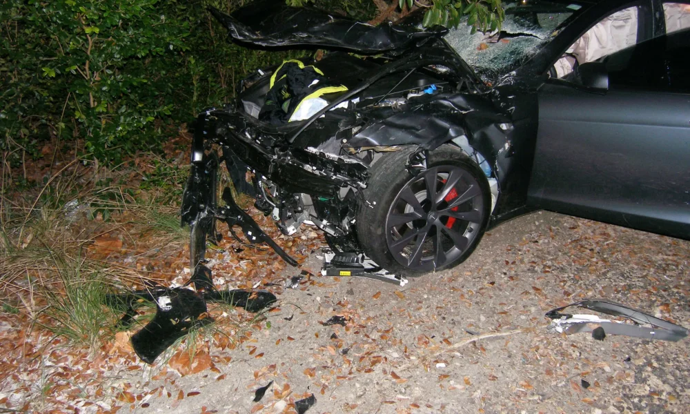Crashed Tesla Model S with severe front-end damage on a roadside near dense foliage.