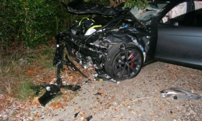 Crashed Tesla Model S with severe front-end damage on a roadside near dense foliage.