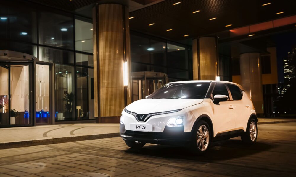 VinFast VF 5 in Brahminy White parked outside a modern glass building at night, LED V-signature DRLs and headlights on, compact city SUV stance with five-spoke wheels.