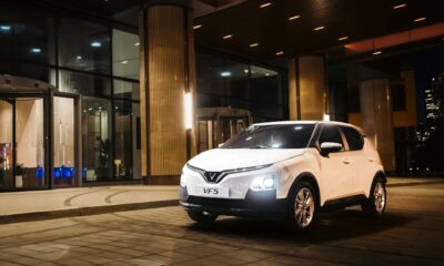 VinFast VF 5 in Brahminy White parked outside a modern glass building at night, LED V-signature DRLs and headlights on, compact city SUV stance with five-spoke wheels.