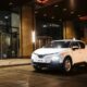 VinFast VF 5 in Brahminy White parked outside a modern glass building at night, LED V-signature DRLs and headlights on, compact city SUV stance with five-spoke wheels.