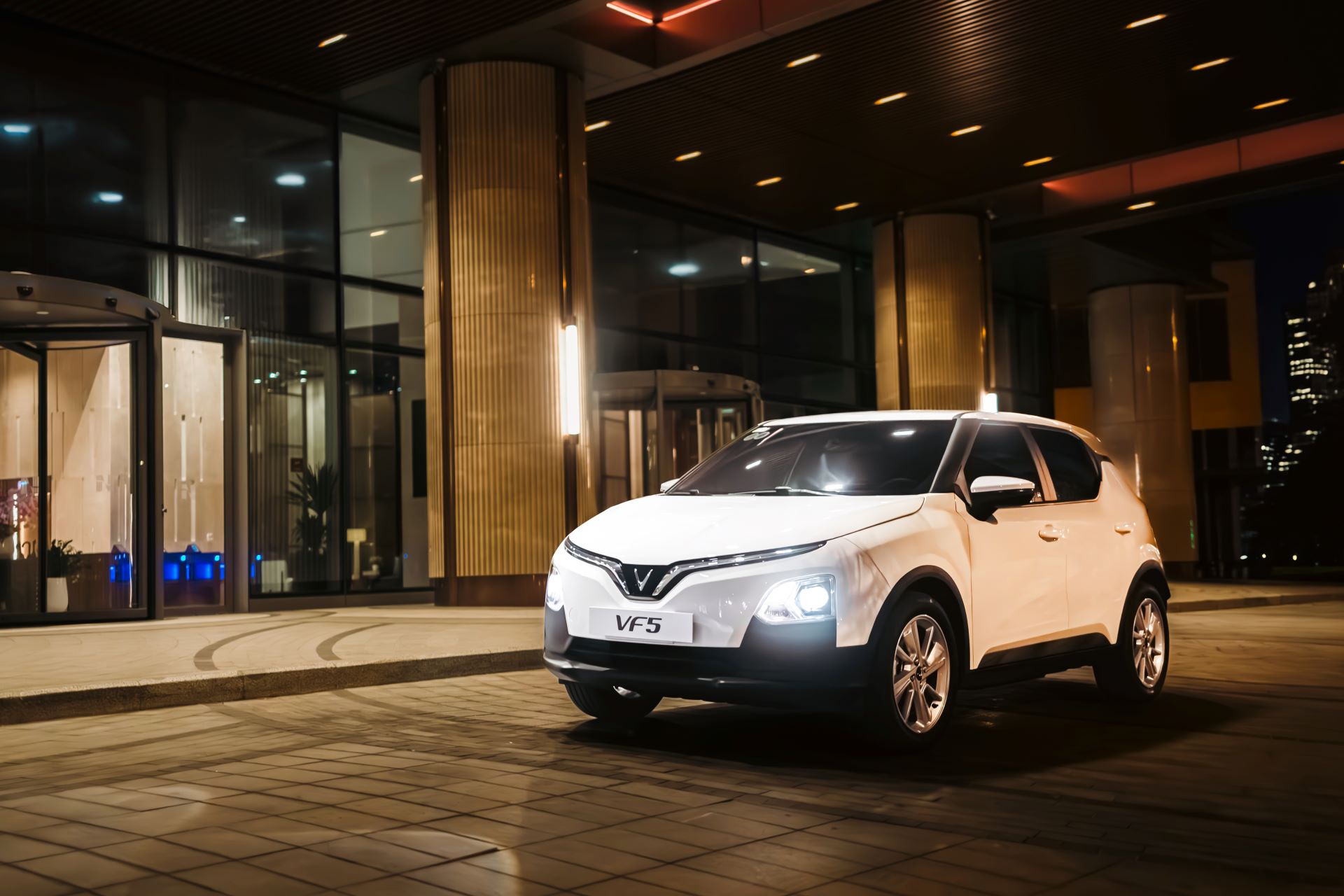 VinFast VF 5 in Brahminy White parked outside a modern glass building at night, LED V-signature DRLs and headlights on, compact city SUV stance with five-spoke wheels.