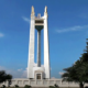 A clear daytime view of the Quezon Memorial Circle monument in Quezon City, framed by trees and a bright blue sky.