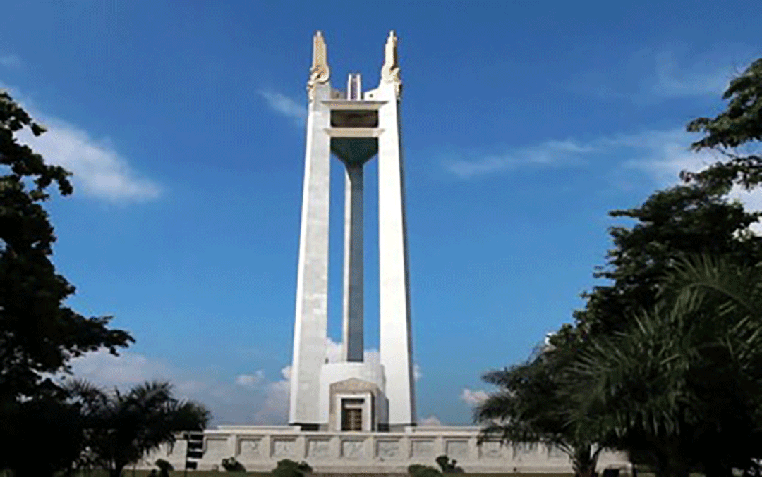 A clear daytime view of the Quezon Memorial Circle monument in Quezon City, framed by trees and a bright blue sky.
