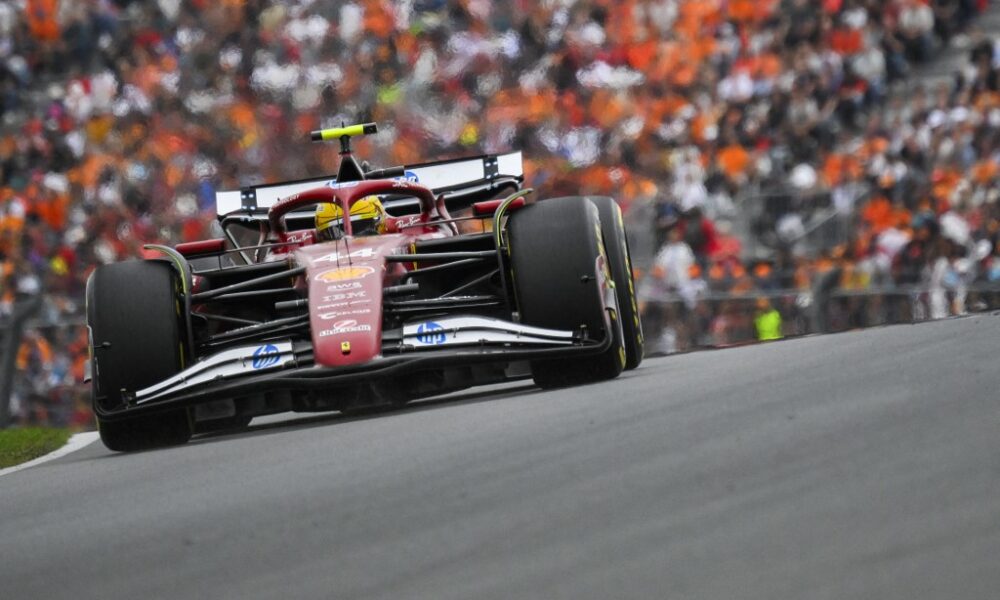 Lewis Hamilton driving his Ferrari F1 car during a race weekend, with a packed grandstand of fans in the background.