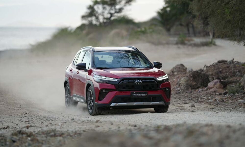 A red Toyota Corolla Cross GR-S HEV driving on a dusty off-road path with trees in the background.