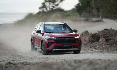 A red Toyota Corolla Cross GR-S HEV driving on a dusty off-road path with trees in the background.