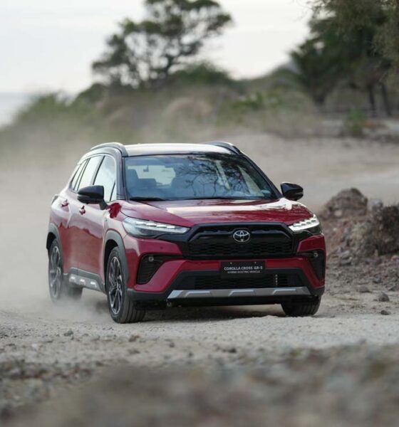 A red Toyota Corolla Cross GR-S HEV driving on a dusty off-road path with trees in the background.