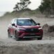 A red Toyota Corolla Cross GR-S HEV driving on a dusty off-road path with trees in the background.