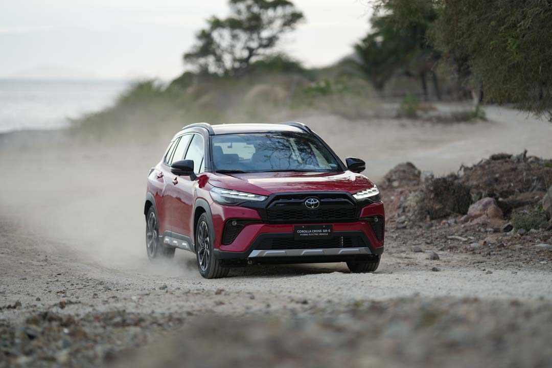 A red Toyota Corolla Cross GR-S HEV driving on a dusty off-road path with trees in the background.