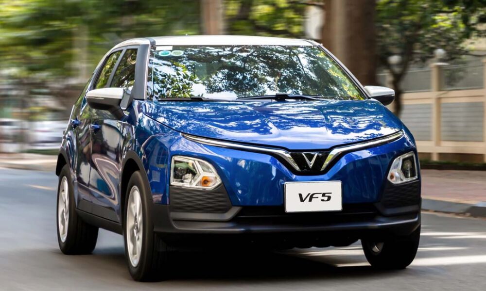 Blue VinFast VF 5 electric crossover driving on a city street with trees and buildings in the background.