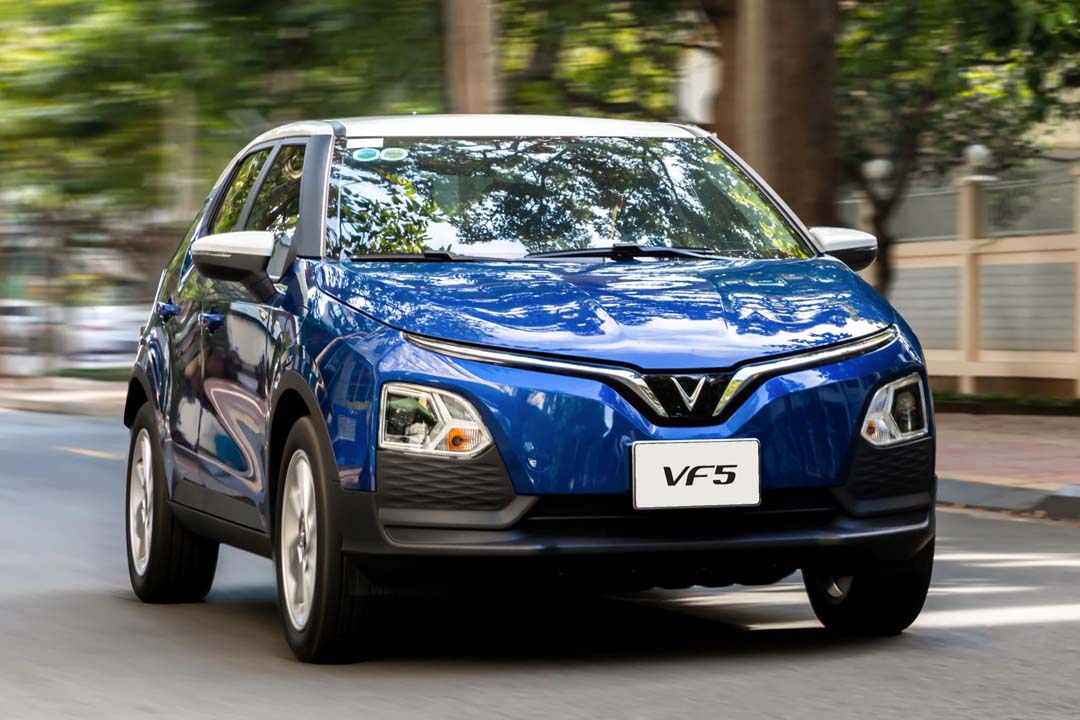 Blue VinFast VF 5 electric crossover driving on a city street with trees and buildings in the background.