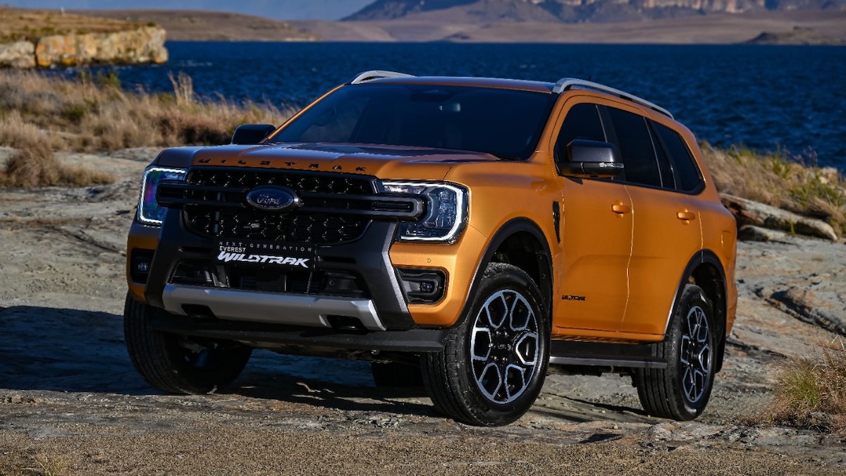 Ford Everest Wildtrak in Luxe Yellow parked on rocky terrain near a lake with mountains in the background.