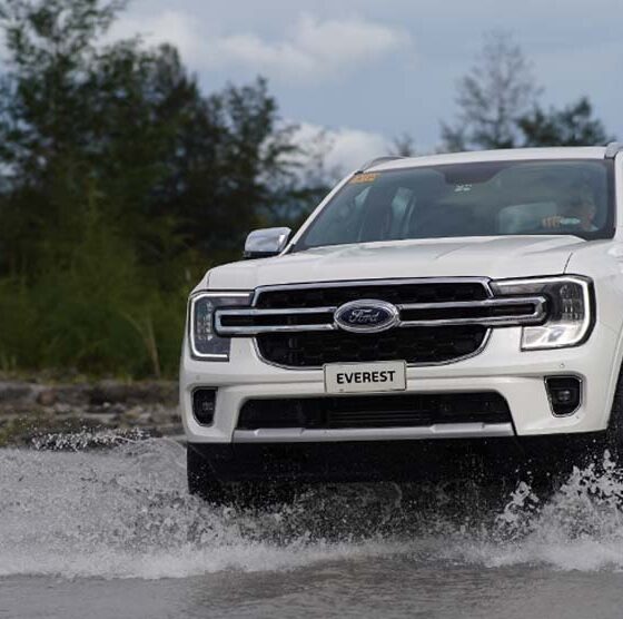 A white Ford Everest drives through shallow water with trees and mountains in the background.
