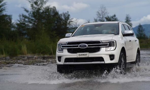 A white Ford Everest drives through shallow water with trees and mountains in the background.
