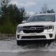 A white Ford Everest drives through shallow water with trees and mountains in the background.