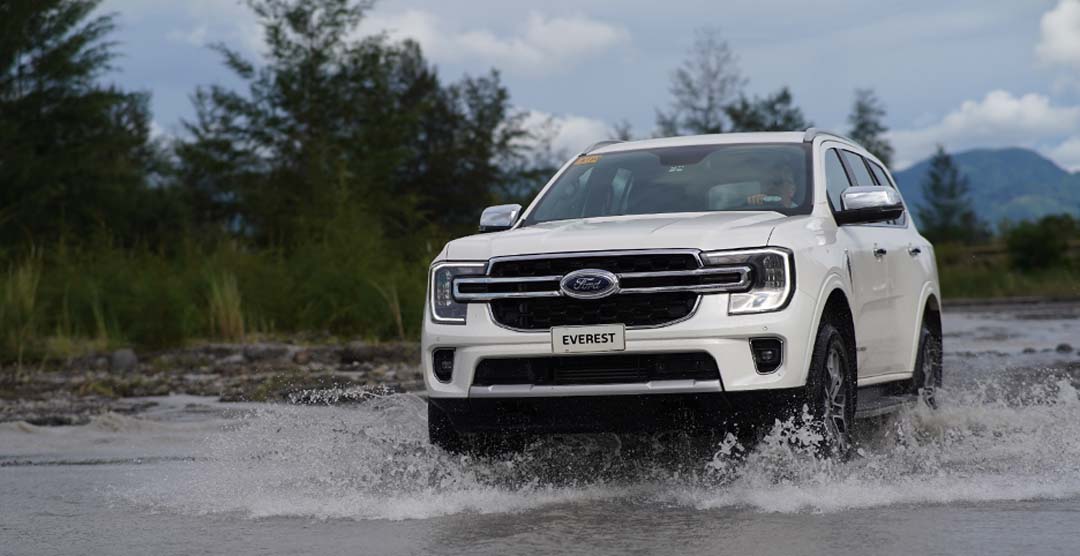 A white Ford Everest drives through shallow water with trees and mountains in the background.